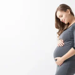 Uśmiechnięta ciężarna kobieta w szarym ubraniu delikatnie dotyka brzucha. To naturalne zmiany, jak rozstępy w ciąży, które są częścią tej wyjątkowej podróży.