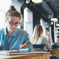 Kobieta w okularach pracuje przy laptopie, pisząc w notatniku. W tle inni pracują, tworząc atmosferę skupienia od dziewiątej do piątej.