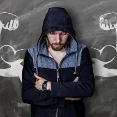 Chłopak w bluzie z kapturem, z ramionami skrzyżowanymi na piersi, stoi przed tablicą z rysunkami bicepsów. Idealny materiał na komplementy dla faceta, który czuje się silny.