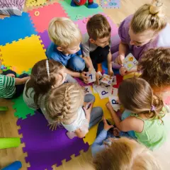Dzieci i dorosła siedzą na kolorowych puzzlach, grając w karty z literami.