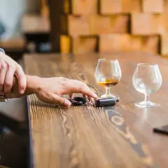 Ręka powstrzymuje drugą przed sięgnięciem po kluczyki do samochodu. Na stole stoją kieliszki z alkoholem, sugerując ryzyko utraty prawa jazdy za alkohol.
