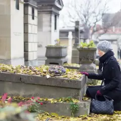 Kobieta klęczy przy grobie, na którym leżą jesienne liście. Przygotowuje się do przeniesienia urny z prochami na inny cmentarz.
