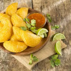 Empanadas colombianas doradas en plato de madera con salsa, limones y cilantro sobre mesa rústica.