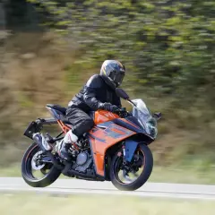 Motociclista con casco y traje negro conduce una KTM RC 390 CV naranja y azul por una carretera con árboles borrosos al fondo.