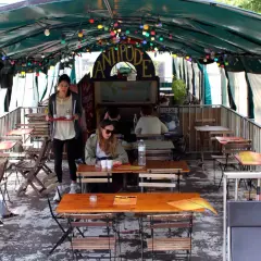 Terrasse animée d'un café-restaurant sur une péniche à Paris, avec des tables en bois, des lumières colorées et des plantes.