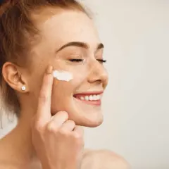Une jeune femme rousse souriante, les yeux fermés, applique de la crème sur sa joue. Elle montre comment appliquer crème visage pour une peau éclatante.