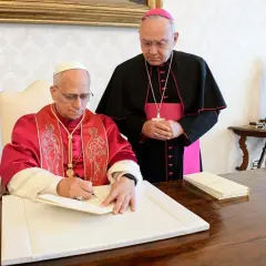 Biskup w purpurowej szacie podpisuje dokument, podczas gdy inny duchowny w czerni i różu stoi obok, obserwując. To ważny moment, być może podpisanie adhortacji.