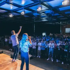 Junge Leute singen auf einer Bühne vor einer großen Menge. Ein Mann hebt die Hand, als ob er eine Botschaft verkündet. Eine moderne Kirche, die urbanes Leben und Gemeinschaft vereint.