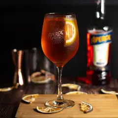 Orzeźwiający drink z aperolem, udekorowany plasterkiem pomarańczy, stoi na drewnianej desce. W tle butelka aperolu i inne akcesoria barmańskie.