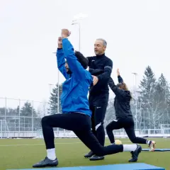 Athletik Training Fußball: Trainer korrigiert Ausfallschritt bei Spieler auf Kunstrasen. Eine Spielerin macht mit.
