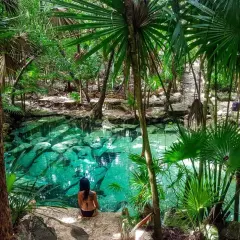 Una mujer se sienta junto a un cenote de agua cristalina en la selva de México, rodeada de exuberante vegetación y palmeras.
