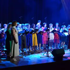 Chór śpiewa pod budą piosenki alfabetycznie. Występują kobiety w kolorowych sukienkach, trzymając nuty.