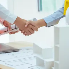 Uścisk dłoni po udanej analizie umowy deweloperskiej. Na stole plany, laptop i makieta budynku.