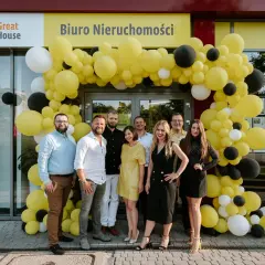 Grupa ludzi pozuje przed biurem nieruchomości ozdobionym żółtymi balonami.