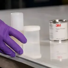 Ręka w fioletowej rękawiczce przygotowuje powierzchnię, obok stoi puszka z primerem 3M Tape Primer 94.