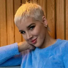 Kobieta z krótkimi blond włosami w niebieskim swetrze opiera głowę na dłoni.