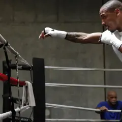 Un boxeur en plein shadow boxing, poings bandés, s'entraîne dans un ring. Un homme en arrière-plan observe.