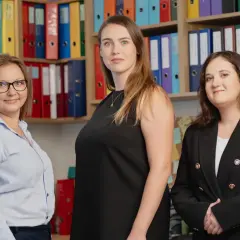 Trzy kobiety w biurze, na półkach widoczne kolorowe segregatory.