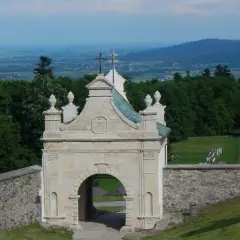 Kamienny łuk z krzyżem na szczycie, otoczony zielenią i widokiem na dolinę.
