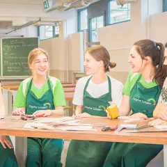 Fünf junge Menschen in grünen Schürzen lachen und arbeiten an einem Holztisch. Hier beginnt ihre kostenfreie Ausbildung Ergotherapie.