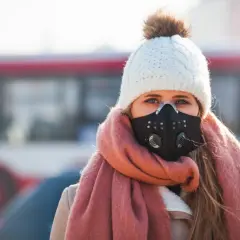 Kobieta w masce antysmogowej, czapce i szaliku na tle miasta.