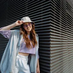 Chica vsco con sombrero y gafas de sol moradas, sonriendo y apoyada en una pared de lamas grises.