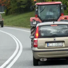 Złoty Fiat Panda jedzie za czerwonym traktorem. Czy można wyprzedzić traktor na podwójnej ciągłej? Droga kręta, drzewa po bokach.