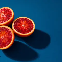 Vibrant fruit photography: three blood oranges halved, revealing their deep red flesh, with a few juice droplets on a deep blue background.