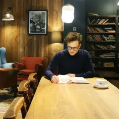 Mężczyzna po francusku, zanurzony w lekturze przy kawie, w przytulnej kawiarni z drewnianymi ścianami i regałami pełnymi książek.