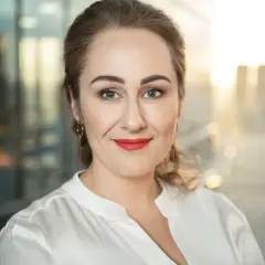 Portret kobiety z rudymi włosami, uśmiechniętej, w białej bluzce. W tle rozmyte miasto.