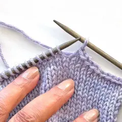 Close-up of hands sewing shoulder seams in knitting with lilac yarn and wooden needles.