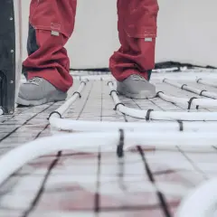 Pracownik w czerwonych spodniach i szarych butach stoi na siatce z rurkami ogrzewania podłogowego. Rozpoczyna się wygrzewanie posadzki po roku.