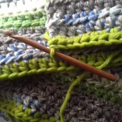 Coperta ai ferri con avanzi di lana, un'opera in corso con un uncinetto di legno che lavora maglie verdi su uno sfondo multicolore.
