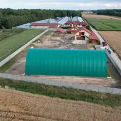 Zielona hala namiotowa na terenie zabudowy siedliskowej, otoczona polami i lasem.