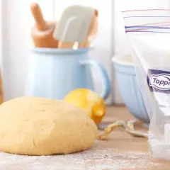 En boll med deg som inte jäst ligger på ett mjölat underlag. Bredvid syns en citron, bakredskap och en fryspåse.