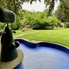 Eine abstrakte schwarze Skulptur steht neben einem blauen Mosaikbecken in einem **Skulpturenpark**. Im Hintergrund ein gepflegter Rasen und Bäume.