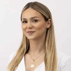 Kobieta z blond włosami i delikatnym makijażem, uśmiecha się lekko, choć jej nienaturalny wyraz twarzy sugeruje, że pozuje do zdjęcia.