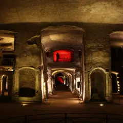 Corridoio sotterraneo illuminato, con nicchie scavate nella roccia e un'apertura rossa in lontananza, evocando l'atmosfera della tomba di San Gennaro.