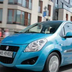 Błękitny Suzuki Splash jedzie ulicą miasta. W tle nowoczesne budynki i ludzie.