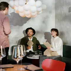 Kelner w różowej koszuli rozmawia z parą przy stoliku. Zastanawiają się, ile zarabia kelner, przeglądając menu.