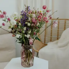 Kolorowy bukiet polnych kwiatów, w tym różowe i białe **gerbery w wazonie**, dodaje uroku przytulnemu wnętrzu z wiklinowym fotelem.
