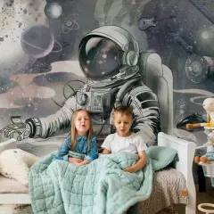 Dzieci siedzą pod kołdrą na łóżku, a za nimi wspaniała tapeta kosmos dla dzieci z astronautą i planetami.