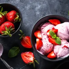 Pyszne lody z truskawek w miseczce, ozdobione świeżymi owocami i miętą. Obok miseczka z całymi truskawkami i gałkownica.