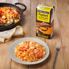 Arroz meloso con gambas y trozos de pescado en paellera y plato, junto a un cartón de fumet.