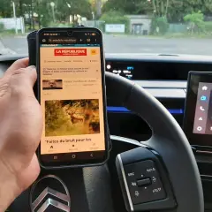 Main tenant un téléphone au volant, risque d'amende. L'écran affiche des articles d'actualité, un danger pour la conduite.
