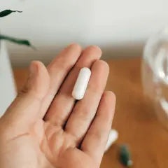 Dłoń trzyma białą kapsułkę. Obok szklanka wody i roślina. Uważaj na nadmiar magnezu objawy, by nie przedawkować.