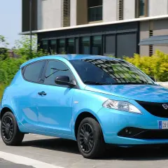 Lancia Ypsilon 1.3 Multijet Diesel azzurra in movimento, con sfondo di edifici moderni e vegetazione.