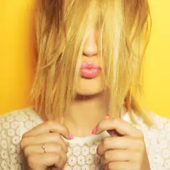 Ein Mund mit rosa Lippen schaut hervor, umgeben von zerzaustem, gelbem Haar. Hände mit pinken Nägeln halten das Haar zurück.