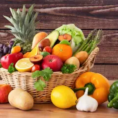 Cesto pieno di frutta e verdura fresca: ananas, uva, mele, arance, limoni, pomodori, peperoni, patate, cetrioli, asparagi e lattuga.