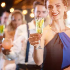 Uśmiechnięta kobieta trzyma kieliszek z drinkiem z prosecco i wódką, w tle bawią się inni goście.